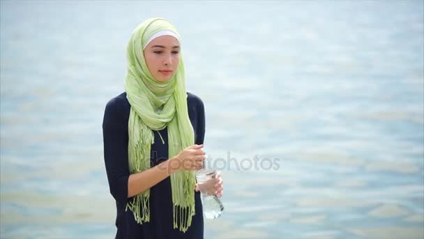 Una joven en un hiyab bebe agua mineral de la botella en un día caluroso al aire libre — Vídeos de Stock