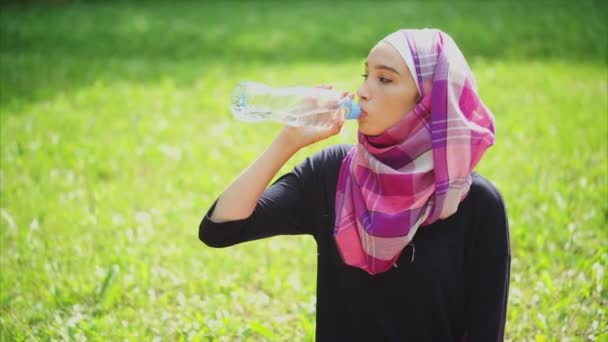 Een Moslimvrouw drinkt mineraal water uit een fles in de parken op een zomerdag — Stockvideo