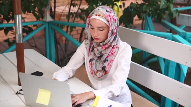 Uma mulher muçulmana moderna em um hijab imprime uma carta a um parceiro de negócios no laptop — Vídeo de Stock