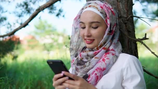 Una mujer con un hijab charlando con amigos en un teléfono inteligente una dama está en el parque — Vídeos de Stock