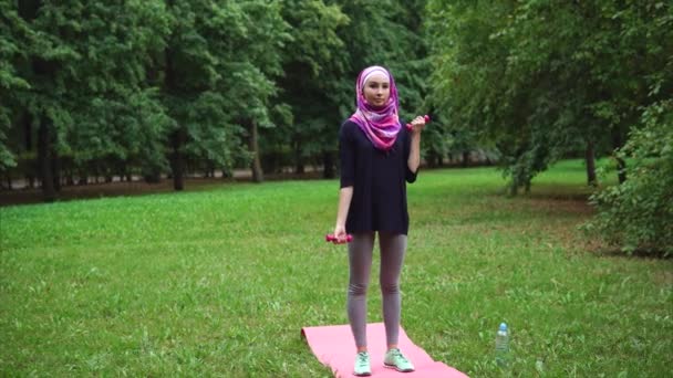 Unga muslimska kvinnan i en burka gör övningar med hantlar i en sommar park — Stockvideo