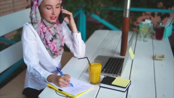 Una mujer musulmana hace una llamada importante en el teléfono móvil toma notas en un cuaderno — Vídeos de Stock