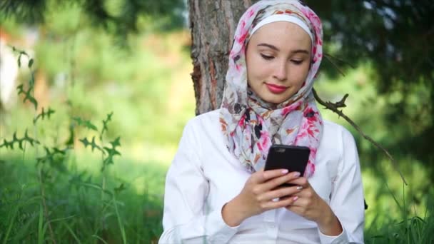 Een moslim in hijab communiceert op de telefoon met vrienden, ze zit in een park — Stockvideo