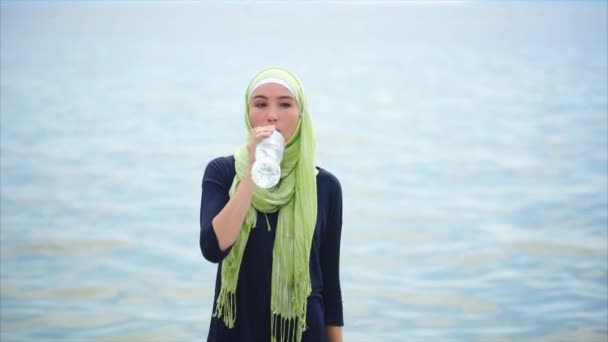 Una joven en un hiyab bebe agua mineral de la botella en un día caluroso al aire libre — Vídeos de Stock