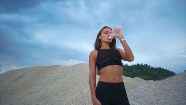 Mujer joven bebe agua sin gas de una botella de plástico en la naturaleza — Vídeos de Stock