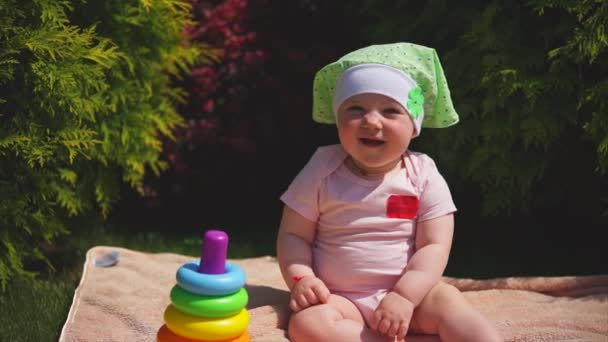 Un recién nacido feliz está sentado en una toalla en el parque, el siguiente es una pirámide de juguetes. — Vídeo de stock
