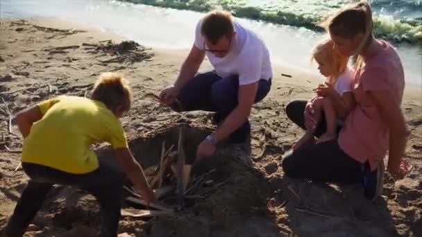 Una familia joven con niños está encendiendo un fuego cerca del mar en un picnic en la playa — Vídeos de Stock