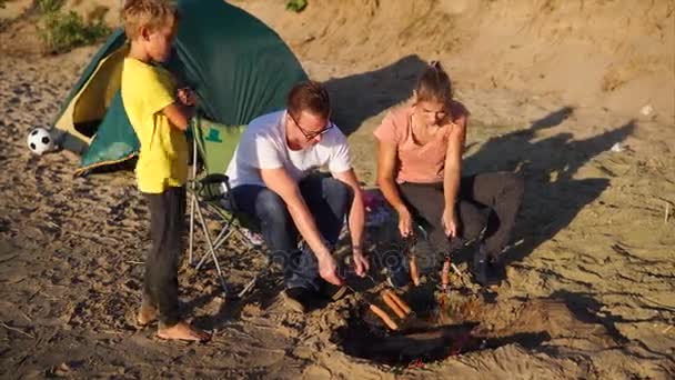 Joven familia fríe salchichas en llamas en la playa en un día de verano ventoso — Vídeos de Stock