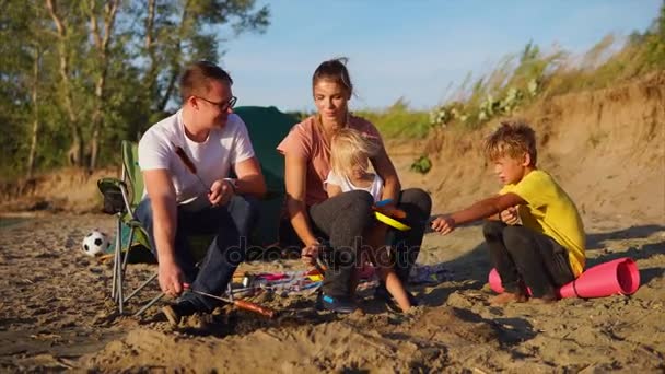 Молодая семья жарит сосиски на шашлыках в огне на пляже в ветреный летний день — стоковое видео