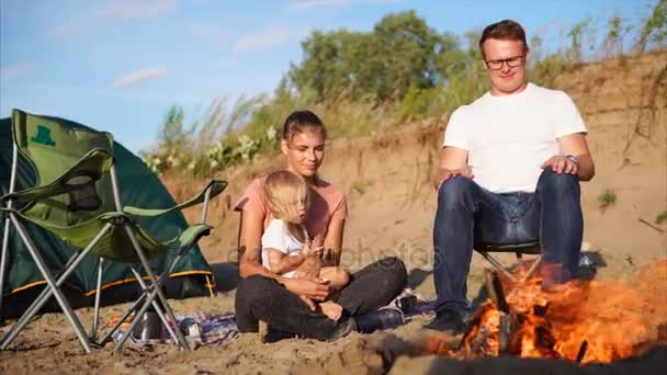 Молодая семья с ребенком, сидящим у костра на природе летом, рядом с палаткой — стоковое видео