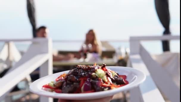 El camarero lleva una ensalada para la pareja que cena en un restaurante cerca del agua — Vídeo de stock