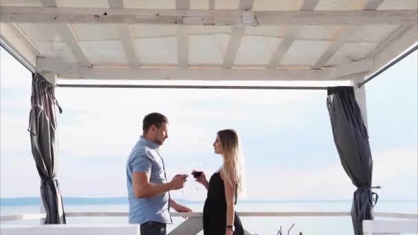 Jóvenes y bonitos cónyuges beben vino de copas en la terraza cerca del mar — Vídeo de stock