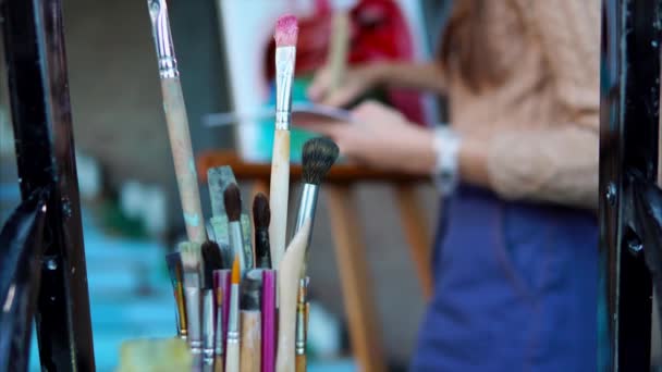 Close up shot of dirty brushes, in the background a woman paints a picture — Stock Video