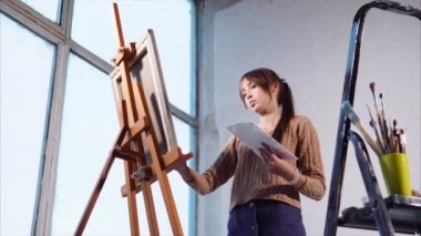 Genç kadın büyük pencereler ile sanat alanda tuval üzerine yağlı boya resim yapıyor.