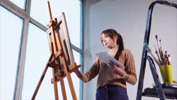 Mujer joven pinta pinturas al óleo sobre lienzo en el espacio de arte con grandes ventanales — Vídeo de stock