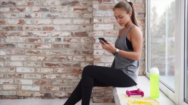 Молодая женщина, которая недавно занялась спортом, проверяет социальные сети в своем телефоне — стоковое видео
