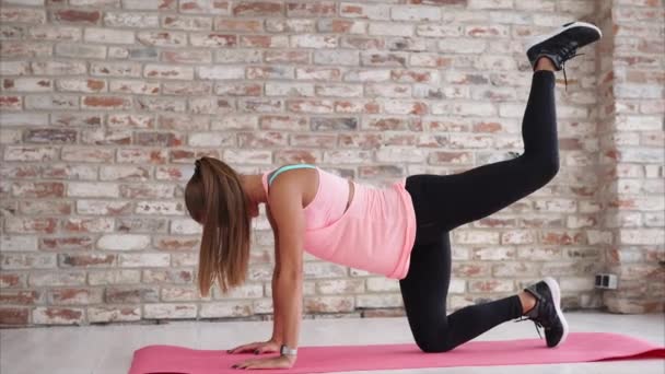 Mujer con el pelo largo y rubio está de pie en una alfombra deportiva en un gimnasio y las piernas en aumento — Vídeos de Stock