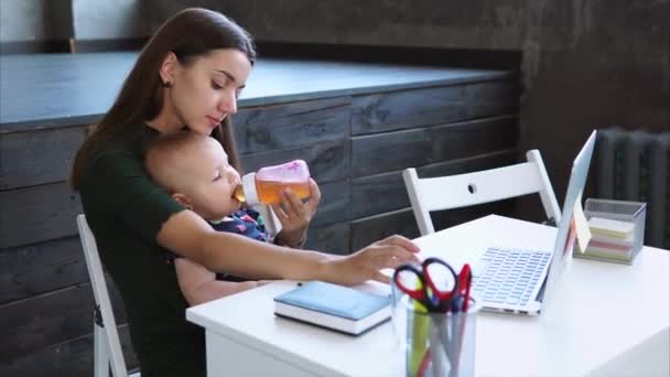Młoda matka trzyma swoje dziecko na ręce i siedzi przy stole, przy użyciu notebooka. — Wideo stockowe