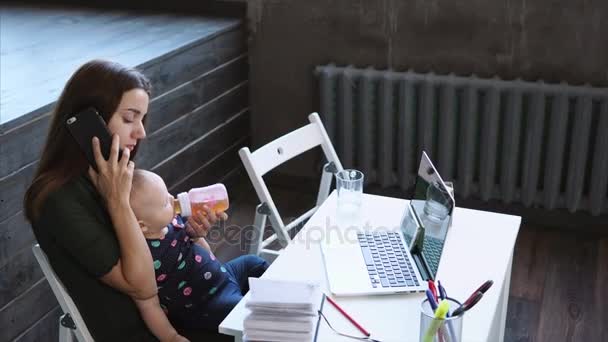 Jonge dame vraagt telefonisch, zittend op een stoel en bedrijf baby op een knieën. — Stockvideo