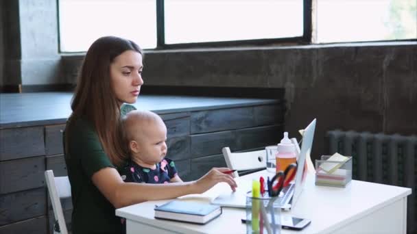 Empresária está fazendo anotações em adesivos, sentado na frente do laptop, com bebê — Vídeo de Stock