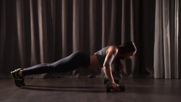 Mladá sportovní žena dělá kliky s činkami na tmavé podlaze v tělocvičně — Stock video