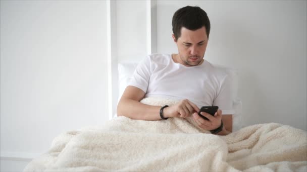 Bonito homem está deitado na cama com smartphone — Vídeo de Stock