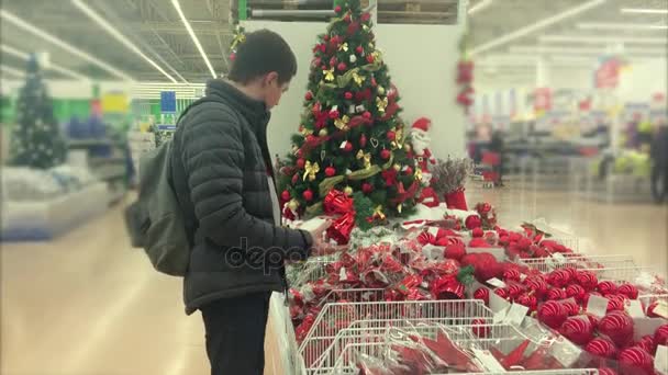 Homem adulto está escolhendo ornamentos para a árvore de Natal em um supermercado — Vídeo de Stock