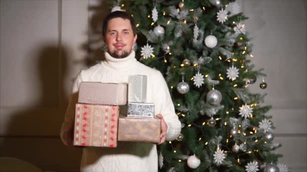 Smiling adult man is standing near Christmas tree in a home and holding gifts — Stock Video