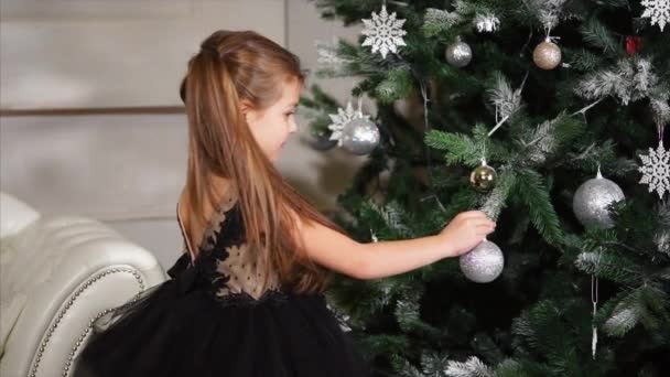 La niña bonita está decorando el árbol de Navidad, se está poniendo una bola de plata . — Vídeo de stock