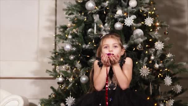 La bambina soffia sui coriandoli scintillanti in mano, contro l'albero di Natale . — Video Stock