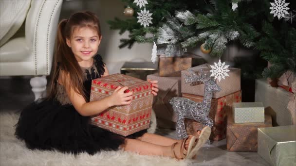 Big-eyed meisje houdt grote doos met de gift van Kerstmis, zittend onder spar — Stockvideo