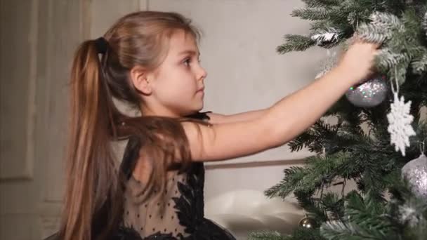 Glückliches Mädchen kleidet vor den Feiertagen einen Weihnachtsbaum, das Baby hält bunte Kugeln — Stockvideo