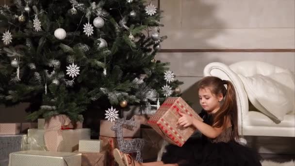 Una niña sosteniendo una caja con un regalo de Navidad ella se sienta en el suelo cerca del árbol de Navidad — Vídeos de Stock