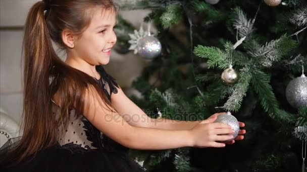 Een klein meisje die in de buurt van de kerstboom zit speelt met een New Years-bal die op een tak weegt — Stockvideo