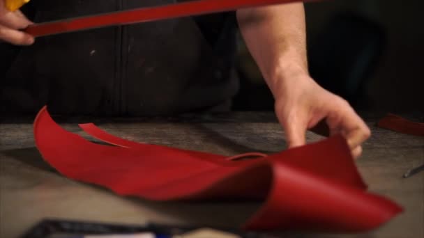 Trabajador de cuero está cortando tiras de cuero pintado rojo en una mesa de trabajo en el estudio — Vídeo de stock