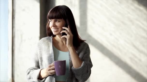Una hermosa dama se sienta cerca de una ventana y bebe té caliente, habla por teléfono — Vídeos de Stock