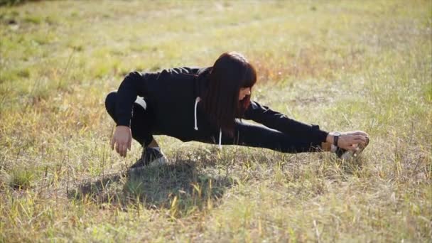 Mujer joven está haciendo ejercicios para estirar las piernas en un campo en un día soleado — Vídeos de Stock
