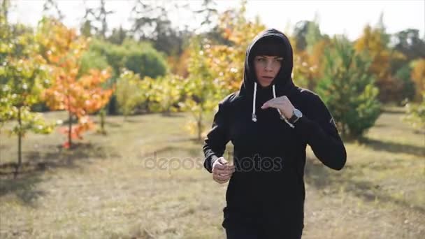 Una mujer con una capucha en la cabeza corre alrededor del parque de otoño durante el día — Vídeos de Stock