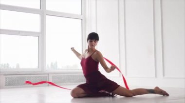 Esmer artistik jimnastikçi bir sınıfta bir katta oturan ve kırmızı kurdele sallayarak
