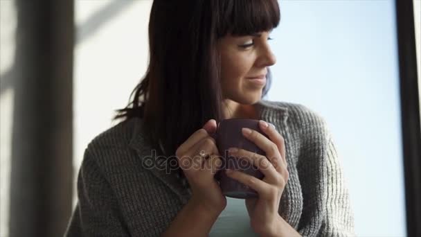 Молодая женщина пьет кофе и смотрит в окно — стоковое видео