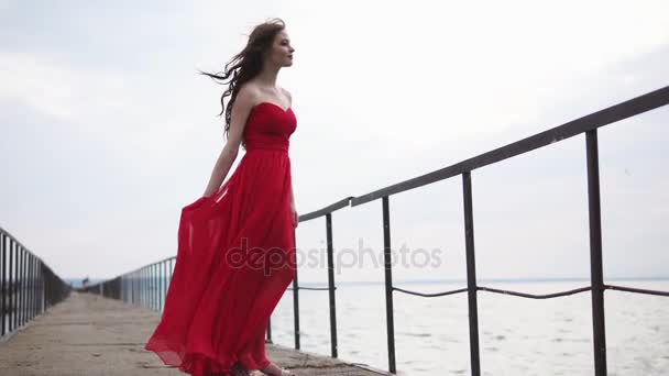 Młoda kobieta w czerwonej sukience stoi na moście w pobliżu morza — Wideo stockowe
