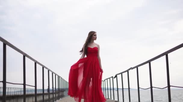 A young and romantic woman dressed in a long red dress and standing on the pier — Stock Video