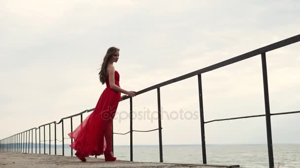 Žena v dlouhých šatech drží na zábradlí u moře, vítr fouká vlasy — Stock video