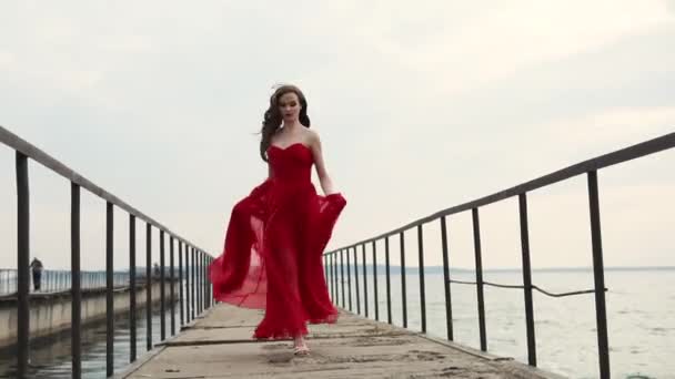Молодая красавица с кудряшками в алом платье бежит по причалу вдоль моря — стоковое видео
