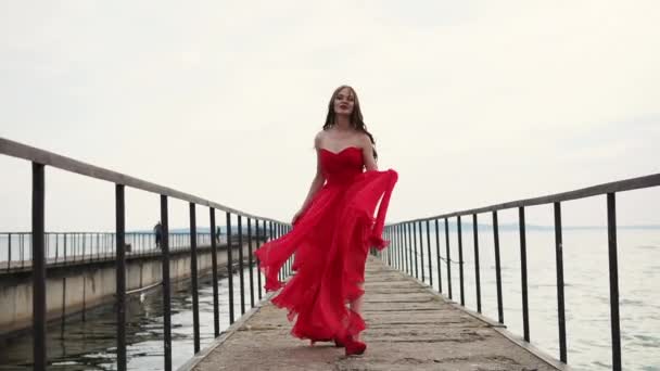 Жінка з кучерявим волоссям прогулюється вздовж пірсу з краєм сукні в руці — стокове відео