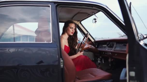 Όμορφη γυναίκα σε ένα κόκκινο φόρεμα κάθεται πίσω από ένα ρολό από ένα σπάνιο αυτοκίνητο ρετρό — Αρχείο Βίντεο