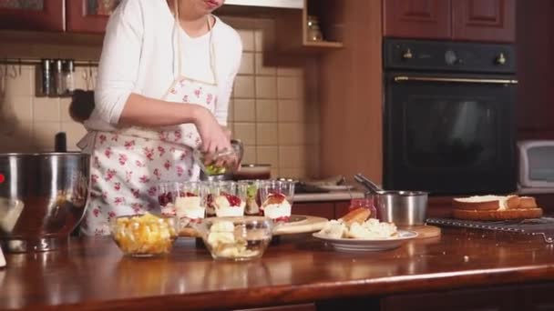 Eine junge Frau in Schürze fügt Kiwi-Stücke in Plastikbecher mit Vanillecreme hinzu — Stockvideo