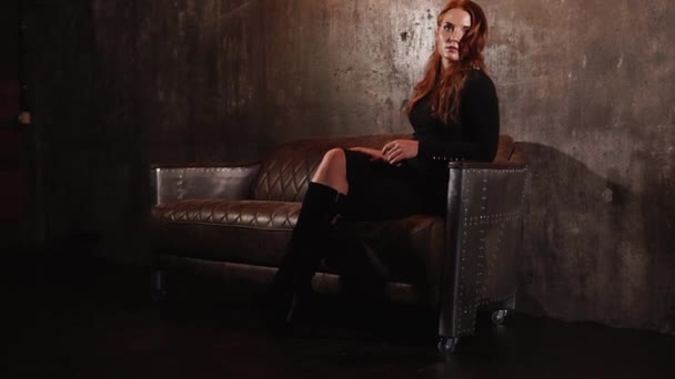 Hermosa mujer de pelo rojo elegante sentado en un sofá de cuero — Vídeo de stock
