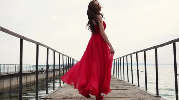 Беззаботная улыбающаяся женщина идет по пристани, размахивая своим шелковым красным платьем — стоковое видео