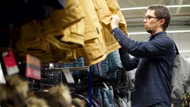 Un hombre adulto elige una chaqueta de invierno, la ropa está en un estante en un centro comercial — Vídeos de Stock
